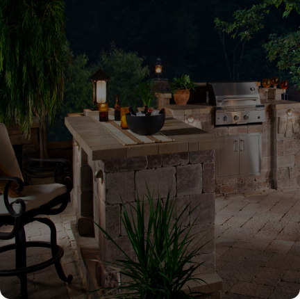 A backyard outdoor kitchen with stone countertops and built-in grill is shown at night. The space is lit by lanterns and features bar stools, potted plants, and various bottles and a bowl on the counter. Trees and foliage provide a natural backdrop.