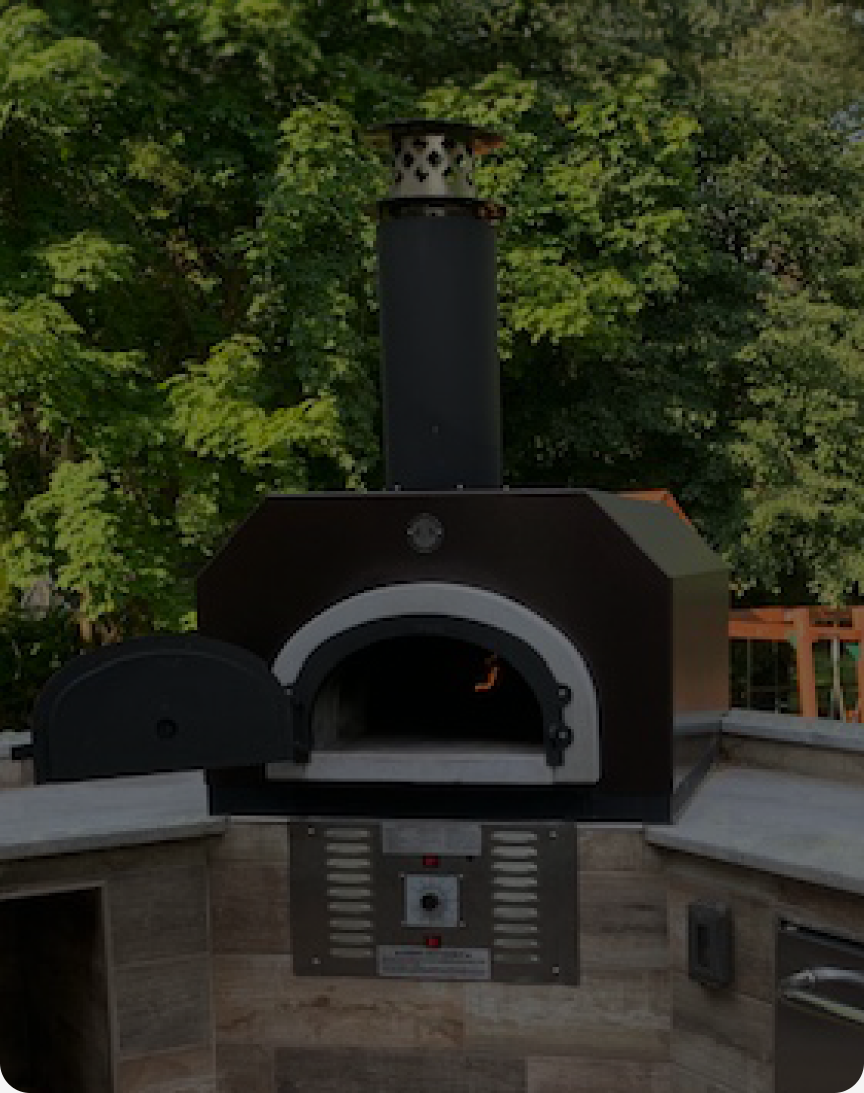 A modern outdoor pizza oven with a stainless steel chimney and an open door, set against a backdrop of lush green trees. The oven is built into a stone countertop, creating an inviting space for outdoor cooking and gatherings.