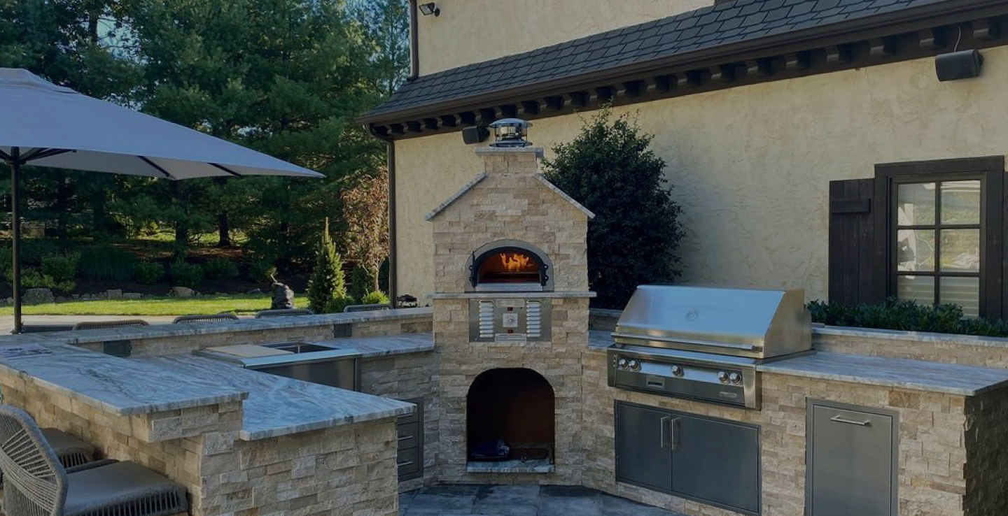 Outdoor kitchen with stone countertops and a built-in grill and pizza oven. The setup includes stainless steel appliances and cabinets with a covered umbrella and a stone wall backdrop. The area is bordered by lush green trees and neatly manicured lawn.