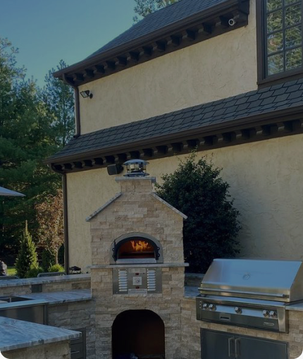 Outdoor kitchen featuring a stone brick pizza oven with a burning fire inside, next to a stainless steel grill. Set against a backdrop of a beige stucco house and surrounded by greenery.