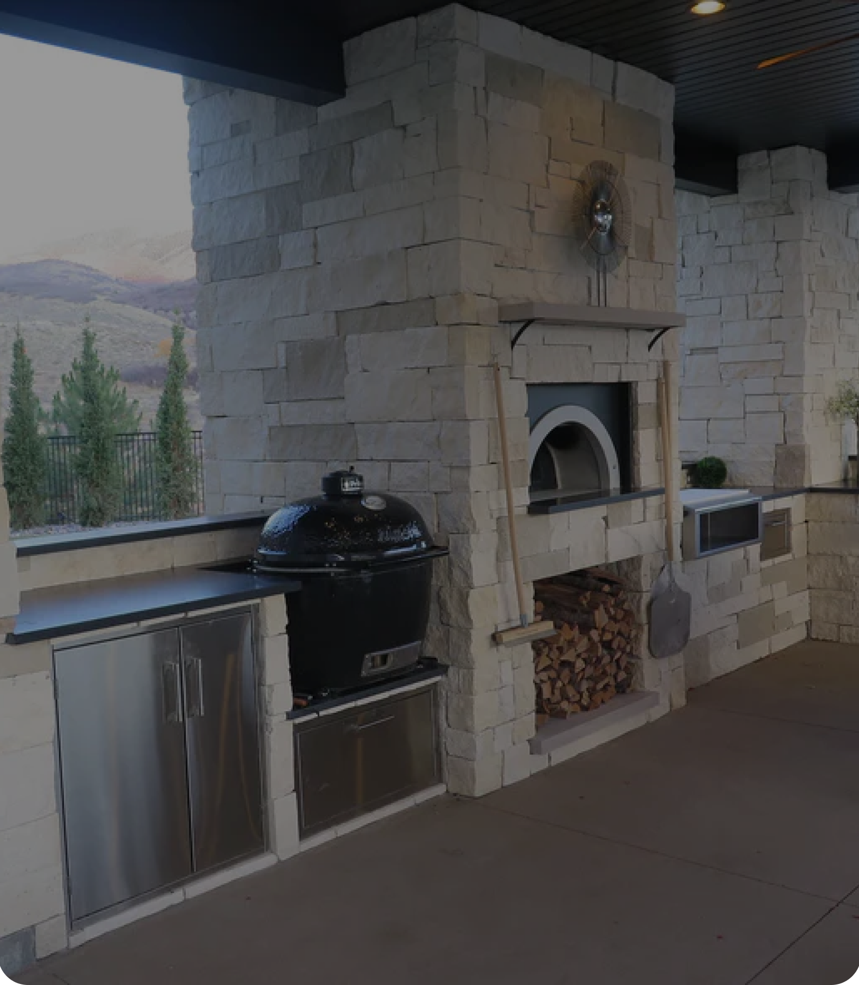 Outdoor kitchen area with a stone wall backdrop, containing a kamado grill, pizza oven, stainless steel cabinets, and a wood storage nook. Tall trees and mountain landscape are visible in the background. A pizza peel and kitchen tools hang on the wall.
