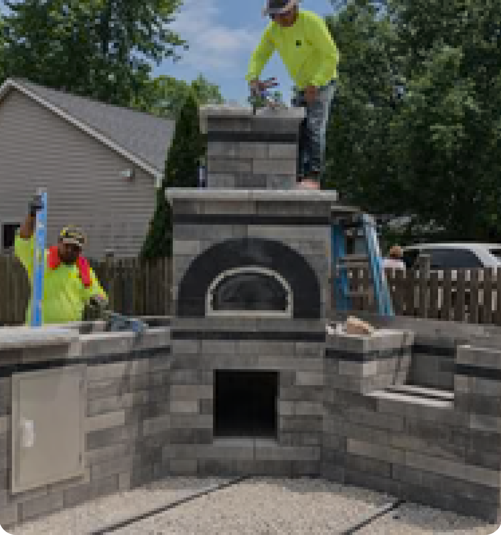 Two construction workers in bright yellow shirts are building an outdoor brick oven and kitchen. One worker is standing on top of the structure working on the chimney, while the other is on the ground leveling a counter. There's a house and trees in the background.