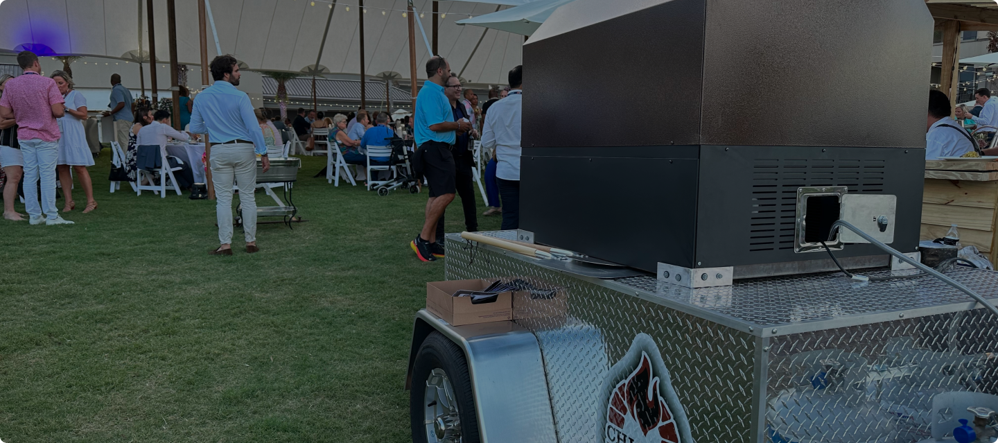A semi-outdoor event with people socializing under a large tent. In the foreground, a metal cart with a box and equipment is visible. Attendees are seated on white chairs and at tables, while some stand and chat on the lawn. String lights are hung across the tent.
