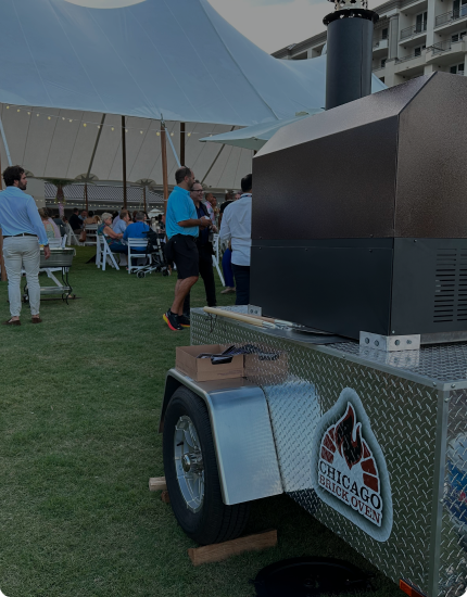 A mobile brick oven with the "Chicago Brick Oven" logo is positioned on a grassy area. People are gathered under a large white tent in the background. The setting appears to be an outdoor event or gathering.