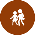 A brown circular icon with white silhouettes of an adult and a child holding hands. Both figures are carrying bags. The adult figure is taller, and the child figure is shorter.