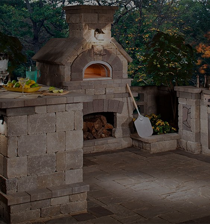 A stone outdoor pizza oven with a wood storage compartment underneath, surrounded by stone countertops and greenery. A pizza peel rests against one side of the oven. The setting is lit by a built-in light and features a table with drinks and candles.