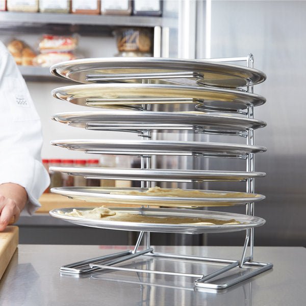 The Chicago Brick Oven 7 Slot Wall Mounted Pizza Pan Rack holds multiple pizza trays stacked vertically, some of which have raw dough placed on them. Positioned on a countertop in a bustling commercial kitchen, the rack stands ready as a person in a white chef's uniform is partially visible in the background.