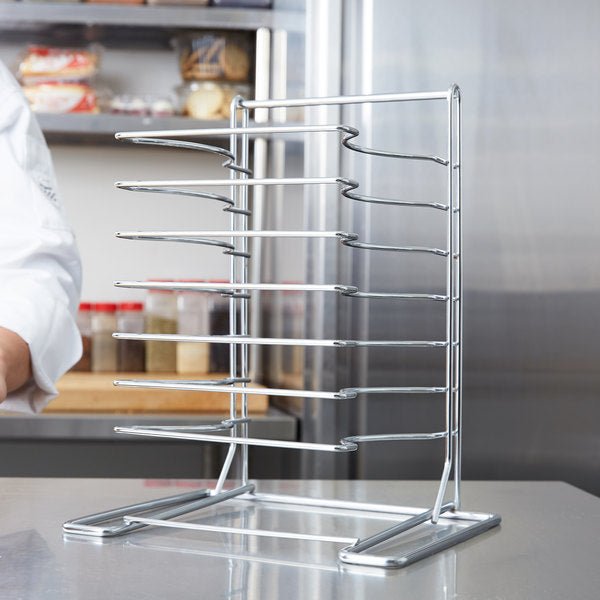 In a bustling commercial kitchen, a 7 Slot Wall Mounted Pizza Pan Rack from Chicago Brick Oven is prominently installed above a gleaming stainless steel countertop. Various food items and containers are blurred in the background, while part of a person in a white coat is partially visible on the left side.