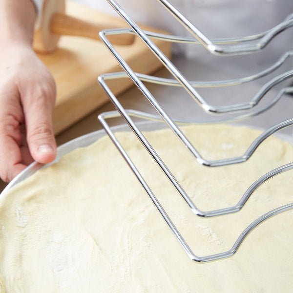 A person is holding a round tray with uncooked dough, poised under the 7 Slot Wall Mounted Pizza Pan Rack by Chicago Brick Oven, commonly found in a commercial kitchen. A wooden rolling pin sits in the background, indicating preparation for baking.