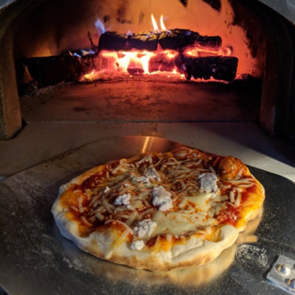 A CBO 500 DIY Kit Wood Fired Pizza Oven from Chicago Brick Oven is in use, with a fire burning in the background. The pizza, topped with tomato sauce, cheese, and pieces of meat, rests on a metal pizza peel inside the oven. The interior glows warmly from the flames against the 27" x 22" cooking surface.