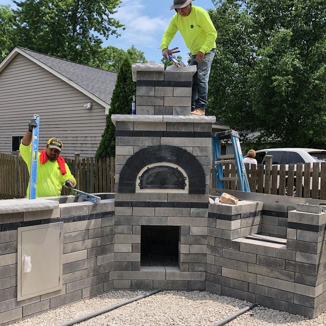 Two workers wearing bright yellow shirts and hats construct a large outdoor brick oven set in a backyard. One stands on top of the structure using tools, while the other measures with a level on the side. A house, trees, and a wooden fence are visible in the background—perfect for enthusiasts of the CBO 500 DIY Kit by Chicago Brick Oven, featuring a 27" x 22" wood-fired pizza cooking surface that combines flexibility with affordability.