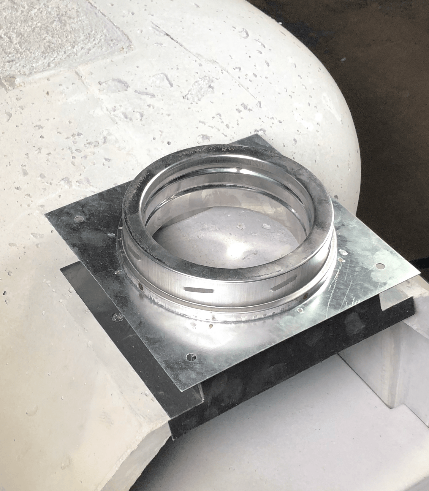 A shiny, steel mechanical flange assembly with a round opening, possibly from the DuraVent 6" Anchor Box for the CBO-750 DIY Kit by Chicago Brick Oven, is mounted on a base plate and positioned on a rough, white surface with visible wear and debris markings. The flange appears to be part of an industrial or mechanical setup and may serve as a replacement part.