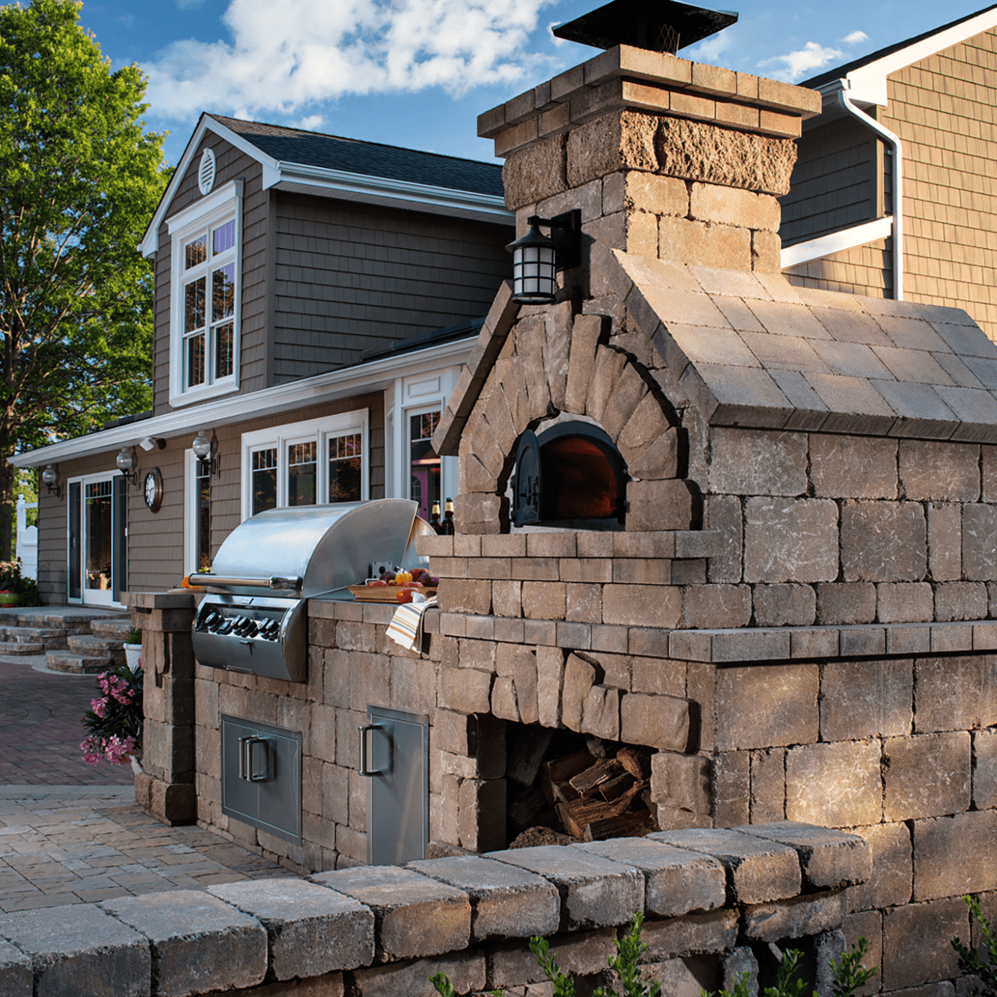 An outdoor kitchen showcases the high-performance CBO 750 DIY Kit, a wood-fired pizza oven by Chicago Brick Oven with a 38" x 28" cooking surface, alongside a gas grill set against a stone patio. Stacked firewood is neatly stored beneath the oven. In the background, there is a modern house with large windows and a gable roof under a partially cloudy sky.