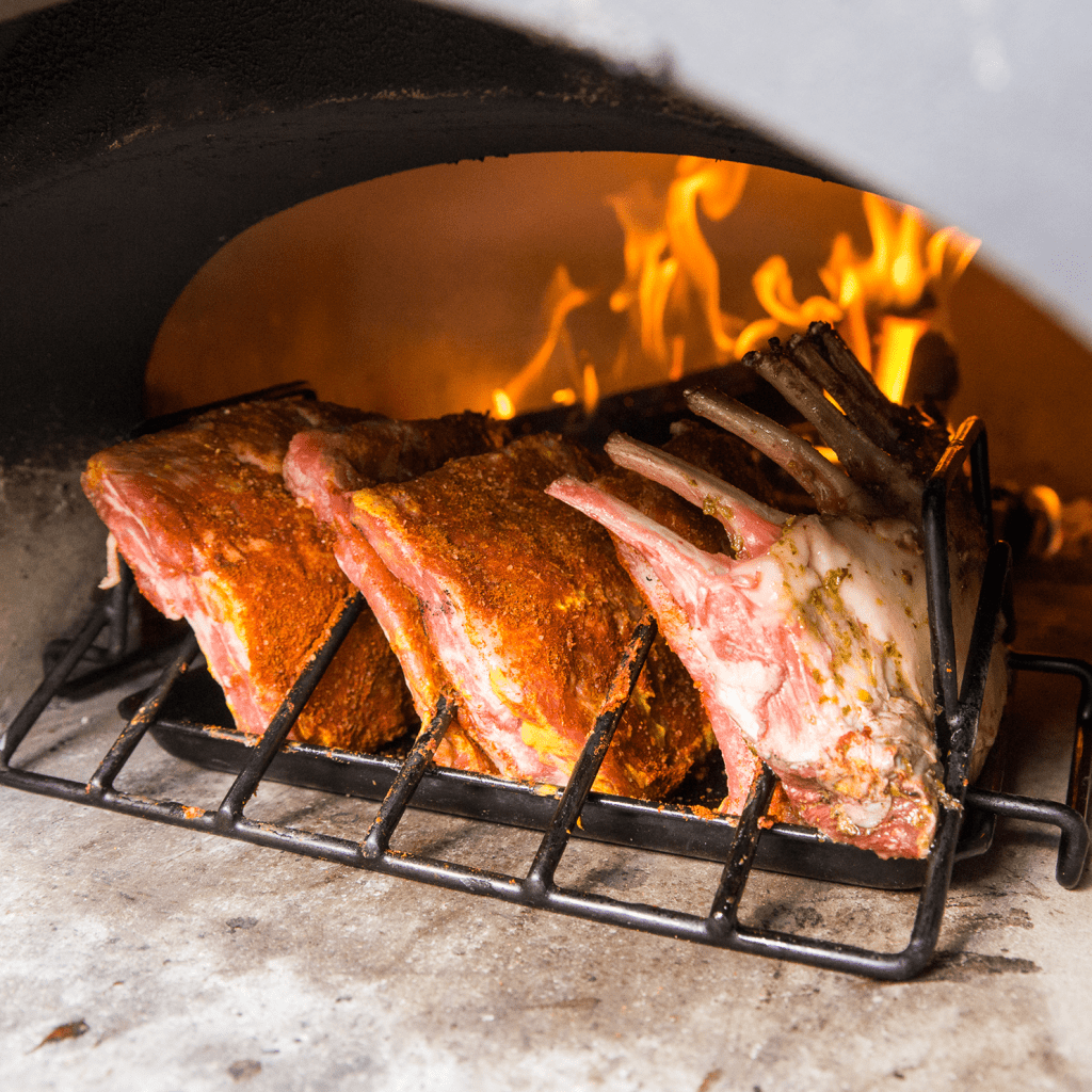 A rack of seasoned lamb ribs sits on a metal grill inside the high-performance CBO 750 DIY Wood Fired Pizza Oven by Chicago Brick Oven. Visible flames and glowing embers indicate high heat, while the ribs are halfway cooked, showcasing a mix of raw pink meat and browned edges on the 38" x 28" cooking surface.