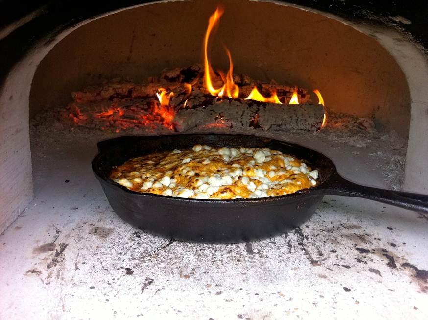 A cast iron skillet filled with a cheesy dish is baking inside a Chicago Brick Oven CBO 750 Hybrid Countertop (Residential) No SKIRT. The flames and burning embers are visible at the back of the wood-fired oven, providing intense heat that is perfectly cooking the food.