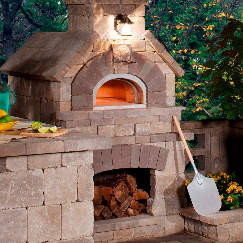 A high-performance Chicago Brick Oven CBO 1000 DIY Kit wood-fired pizza oven, featuring a spacious 53" x 39" cooking surface and an integrated wood storage area below, sits impressively with a metal pizza peel leaning against it. The surrounding area includes a counter topped with lime slices and a drink pitcher, all set against lush foliage in the background. The oven is illuminated by a small light above, highlighting its sleek, stone finish.