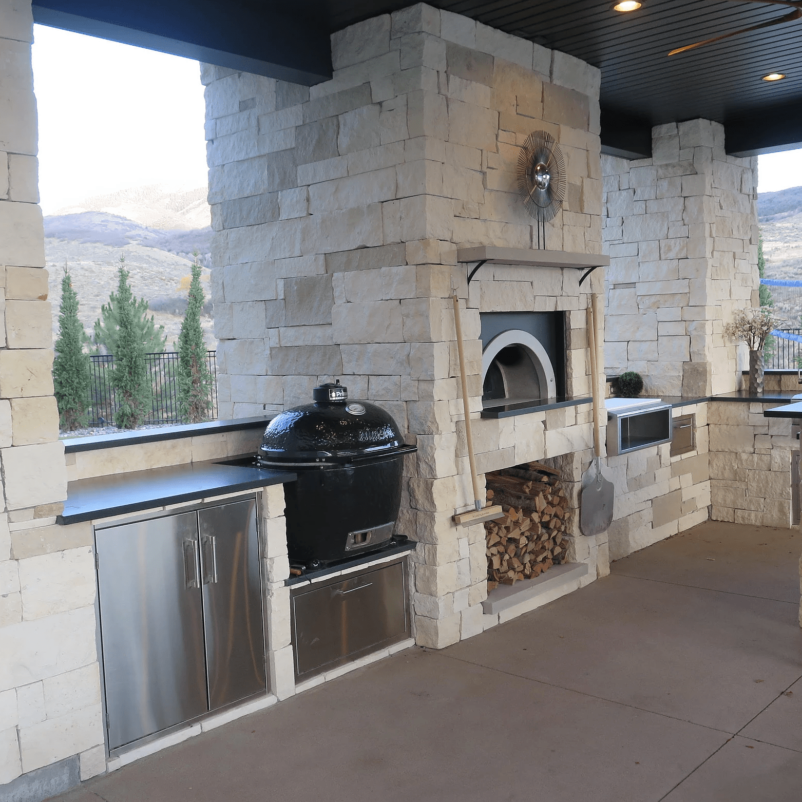An outdoor kitchen with a stone-finished structure features a built-in BBQ grill, outdoor pizza ovens including the Chicago Brick Oven CBO 1000 DIY Kit with a 53" x 39" wood-fired cooking surface, a stainless steel cabinet, and firewood storage. The kitchen overlooks a scenic view of hills and pine trees. There's a ceiling fan and beige flooring.