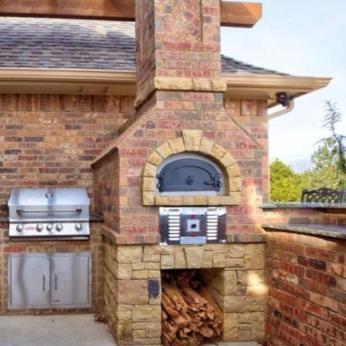 Outdoor brick kitchen featuring the CBO 750 Hybrid DIY Kit (Commercial) from Chicago Brick Oven with a built-in dual-fuel (gas and wood) grill and a wood-burning oven. The grill boasts stainless steel doors beneath it, while the oven, crafted from commercial-grade refractory cement, showcases a curved black door above a firewood storage space. The setup is situated against a brick house under a roof.