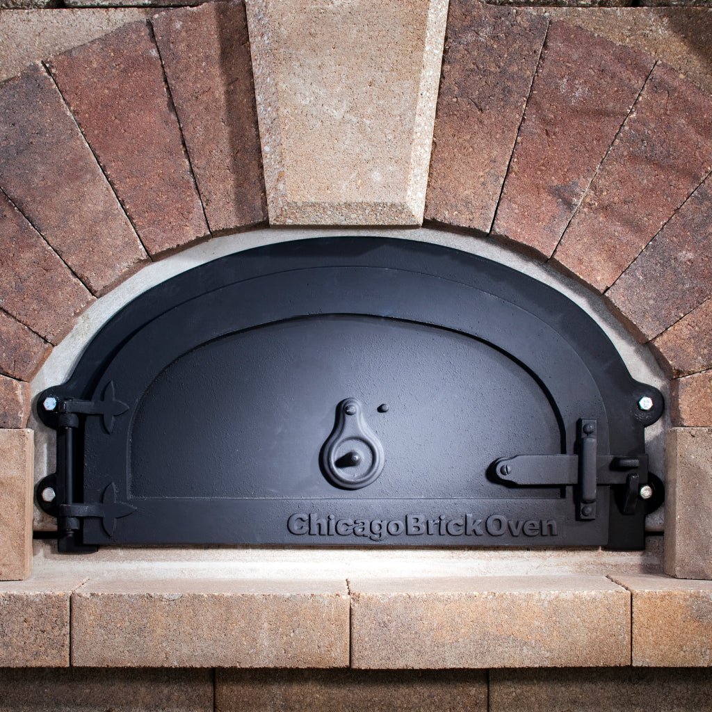 A close-up of a Chicago Brick Oven CBO 500 & 750 Models showcases the oven's distinctive black, arched cast iron door set within a brick facade featuring varying shades of tan and brown stones. The door, which can easily be replaced with a CBO Decorative Door by Chicago Brick Oven, has a simple handle and is framed by the surrounding brickwork.