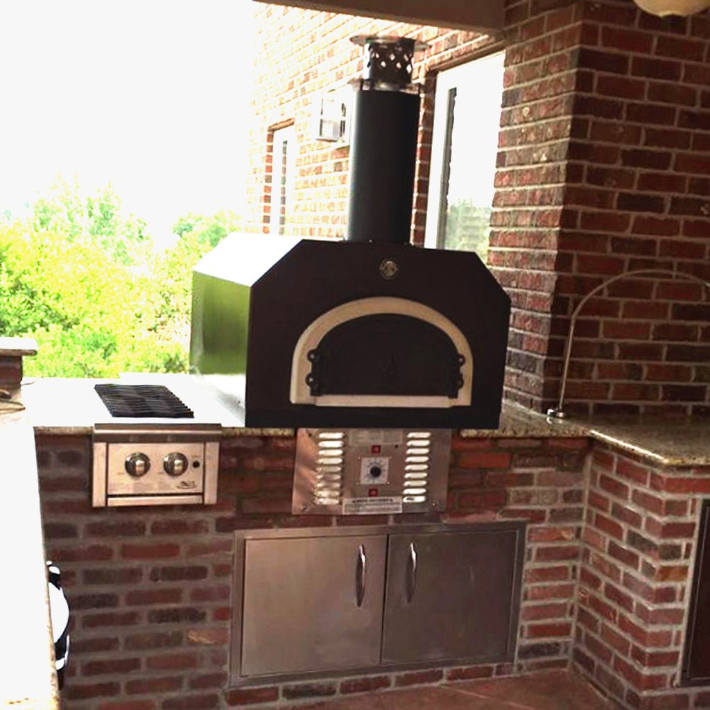 A brick outdoor kitchen featuring a Chicago Brick Oven CBO 750 Hybrid Countertop (Residential) No SKIRT with Dual Fuel (Gas and Wood), a black chimney, a built-in stovetop, and stainless steel storage cabinets. The area is surrounded by brick walls and overlooks a green, leafy landscape.
