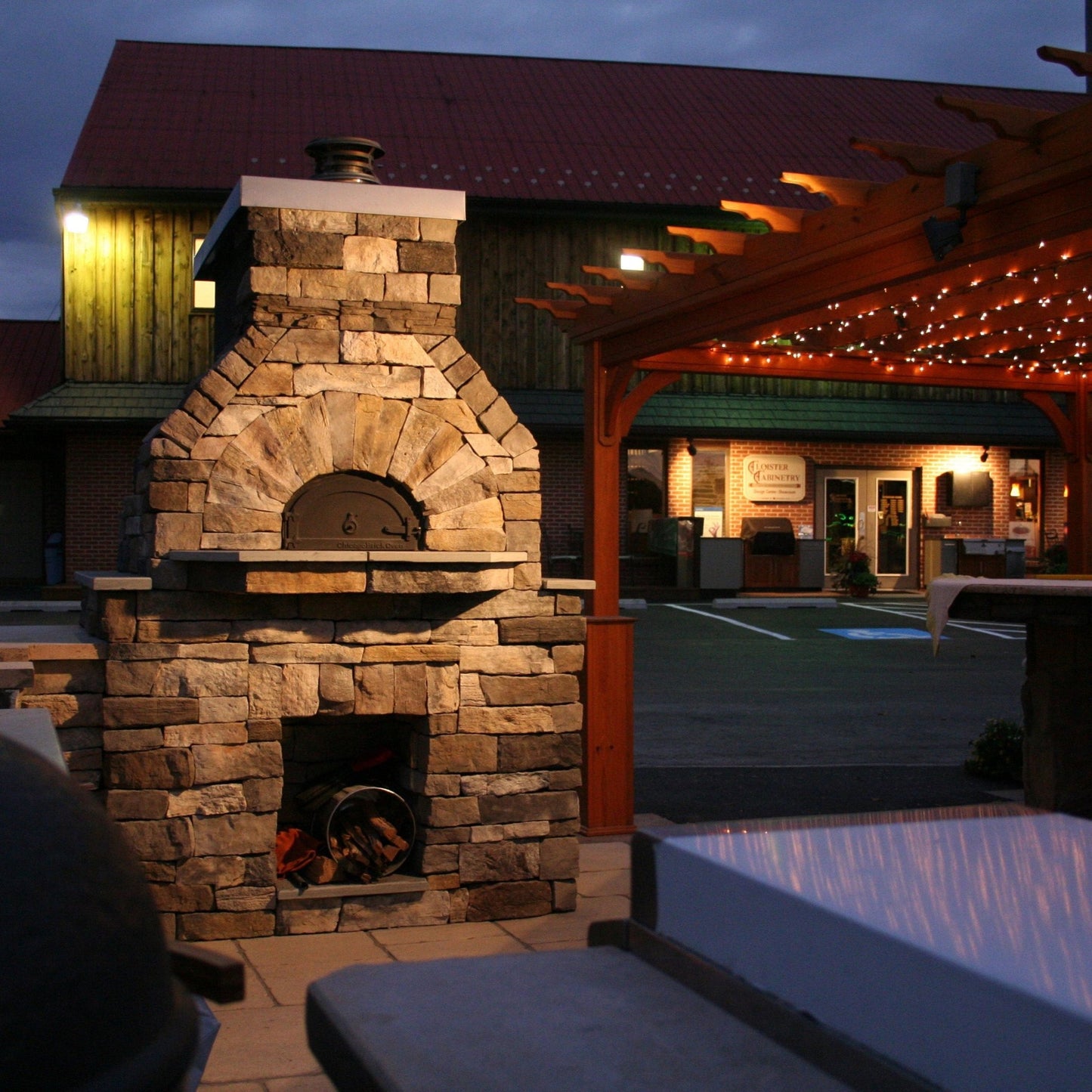 An outdoor courtyard at dusk sets the stage for a cozy, well-lit scene featuring the Chicago Brick Oven CBO 750 DIY Kit wood-fired pizza oven, complete with a stack of firewood. Positioned to the right is a charming wooden pergola adorned with string lights, while various buildings form the backdrop. This inviting atmosphere is warmly enhanced by the high-performance pizza oven's 38" x 28" cooking surface from Chicago Brick Oven's most popular bundle.