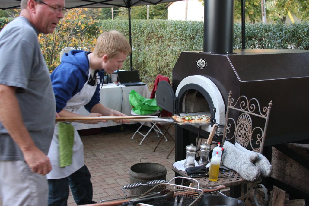 A young man and an older man stand beside a CBO 500 Countertop from Chicago Brick Oven, a wood-fired pizza oven with a 27" x 22" cooking surface. The young man, wearing a white apron, uses a wooden pizza peel to place a pizza into the outdoor oven while the older man observes. The scene is set under a canopy with various cooking tools in the foreground.