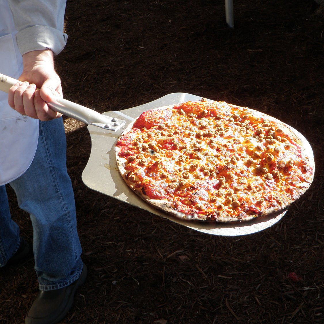 A person holding a freshly baked pizza on a metal pizza peel. The pizza, cooked in a Chicago Brick Oven CBO 750 Hybrid Countertop (Residential) No SKIRT | Dual Fuel (Gas and Wood), is topped with cheese, pepperoni, sausage, and various other toppings. The person is wearing jeans and a white apron. The ground beneath is covered with dark mulch.