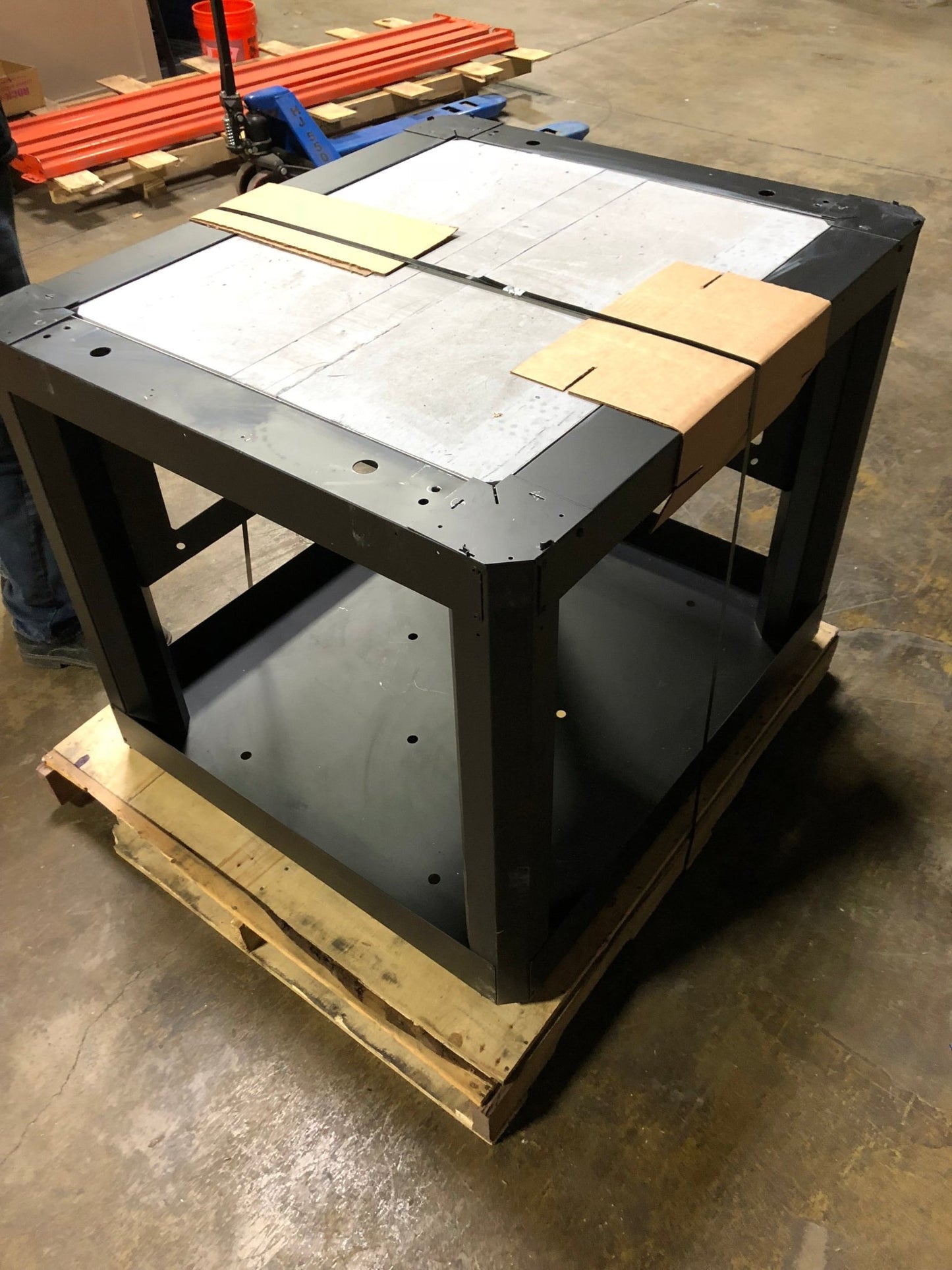 A CBO-750 Mobile Cart / Stand from Chicago Brick Oven, featuring a large black metal frame, is strapped onto a wooden pallet. The stand is partially covered with square, light-colored panels and is positioned on a concrete floor in an industrial setting. Some red pipes and other materials are visible in the background.