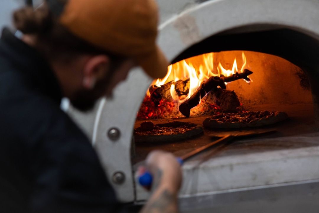 A person in a black shirt and brown cap is using a pizza peel to place pizzas into a Chicago Brick Oven CBO 1000 DIY Kit, with its high-performance wood-fired capabilities and 53" x 39" cooking surface, flames dancing inside creating a warm, cozy atmosphere.