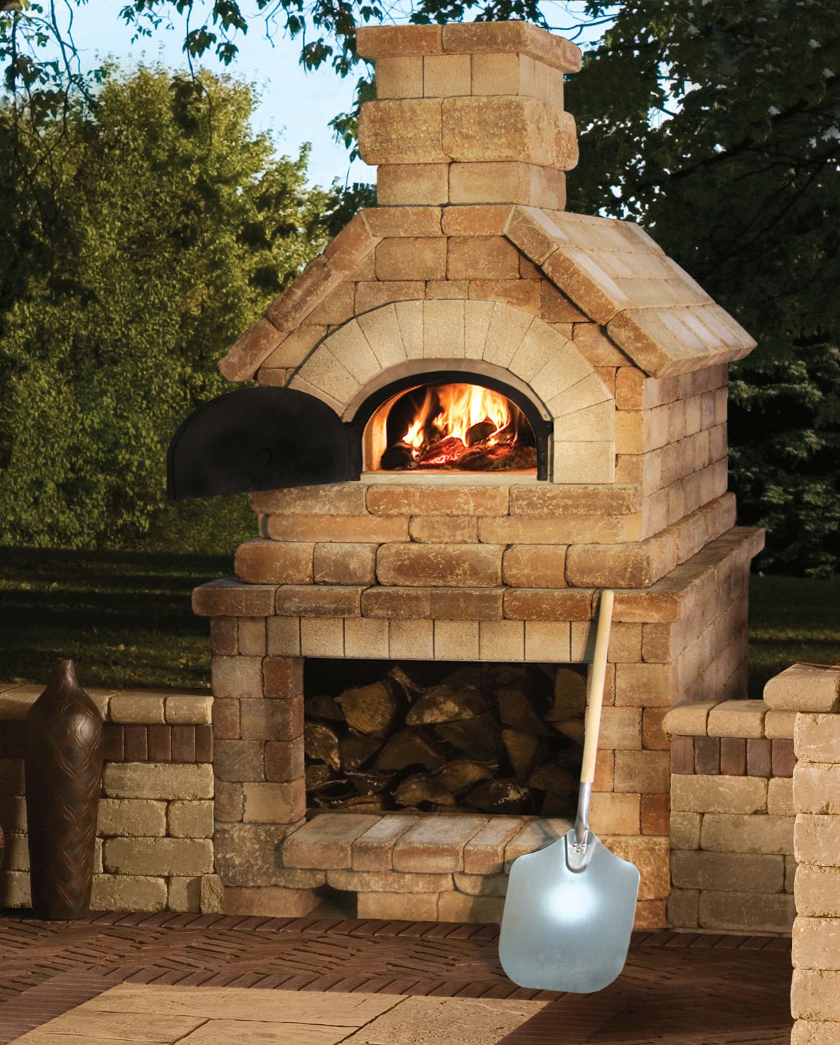 CBO 750 DIY Kit | Wood Fired Pizza Oven by Chicago Brick Oven, our most popular bundle with a 38" x 28" cooking surface, features a high-performance wood-fired oven with a small fire burning inside. Beneath the oven is a storage area filled with chopped wood. A pizza peel leans against the fireplace, and a tree-filled landscape is visible in the background.