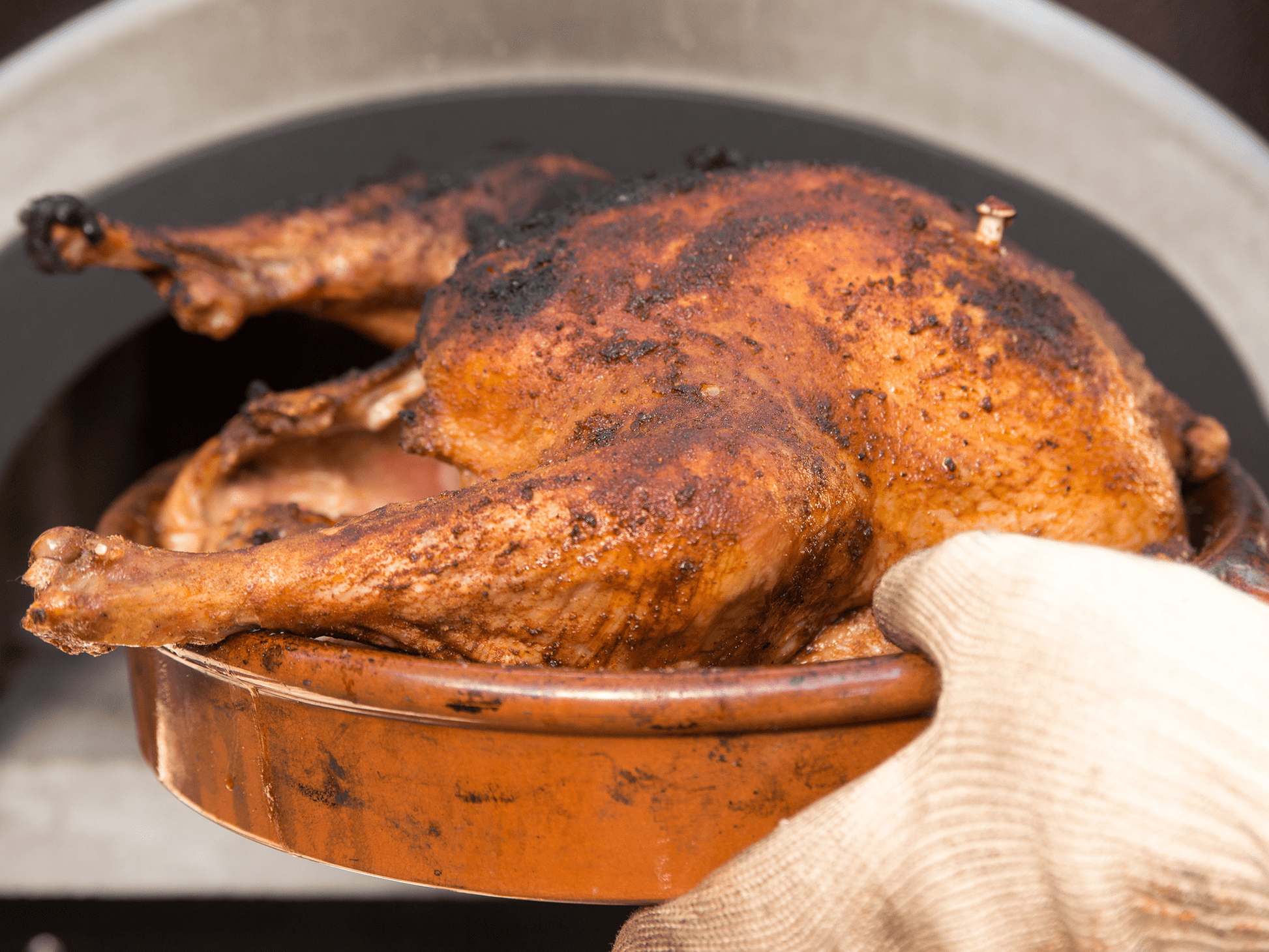 A gloved hand holding a platter with a beautifully roasted whole chicken, well-seasoned and slightly charred, emerging from an oven. The chicken appears golden-brown and crispy, steaming hot and ready to be served, as if cooked in the CBO 750 Hybrid Countertop (Residential) No SKIRT | Dual Fuel (Gas and Wood) by Chicago Brick Oven.