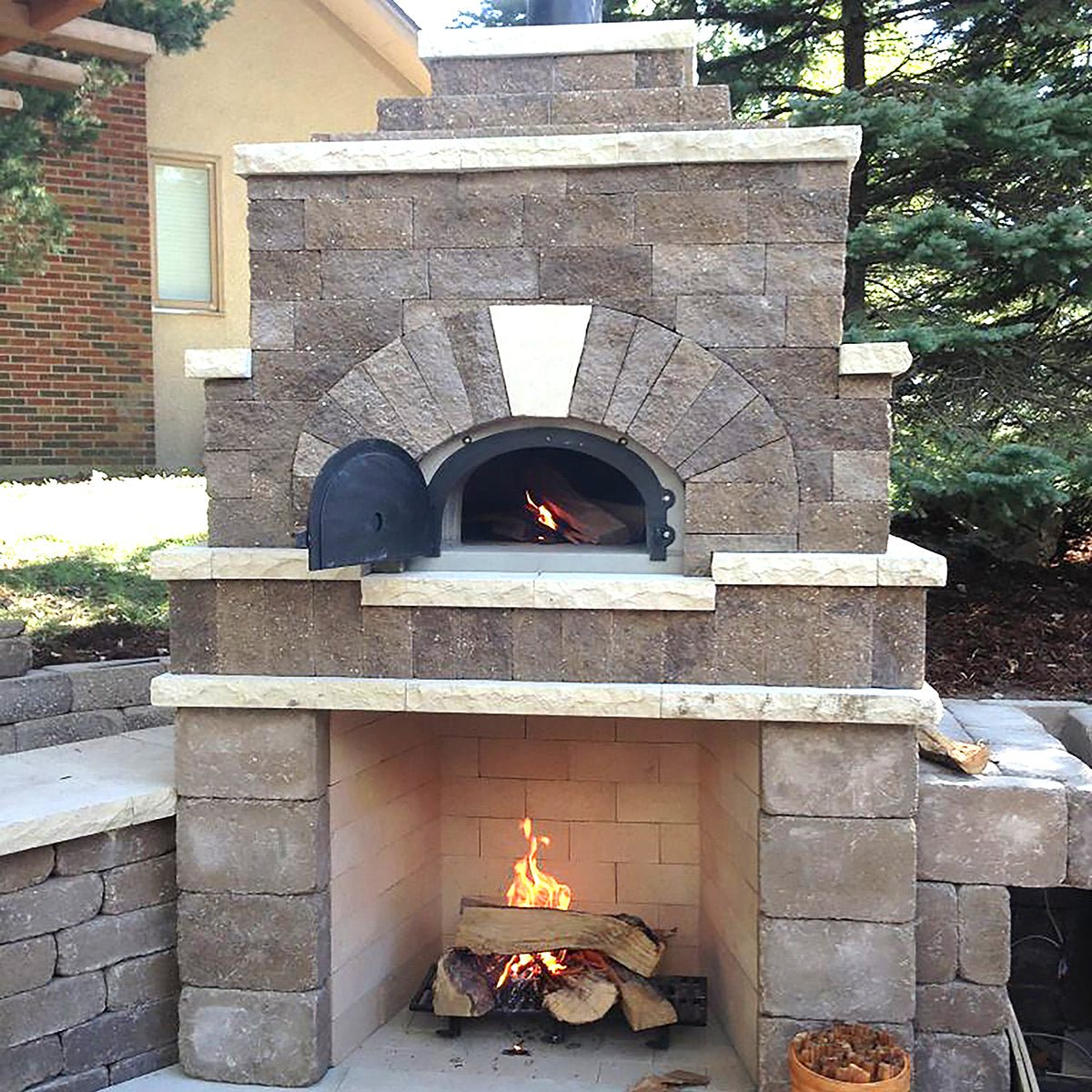 An outdoor two-chambered stone oven with a fire burning in the lower chamber and an access door open to the upper chamber, where flames are also visible. This wood-burning CBO 500 DIY Kit by Chicago Brick Oven, featuring a 27" x 22" cooking surface and offering flexibility alongside affordability, is set against a backdrop of bricks and trees, ideal for baking pizzas.