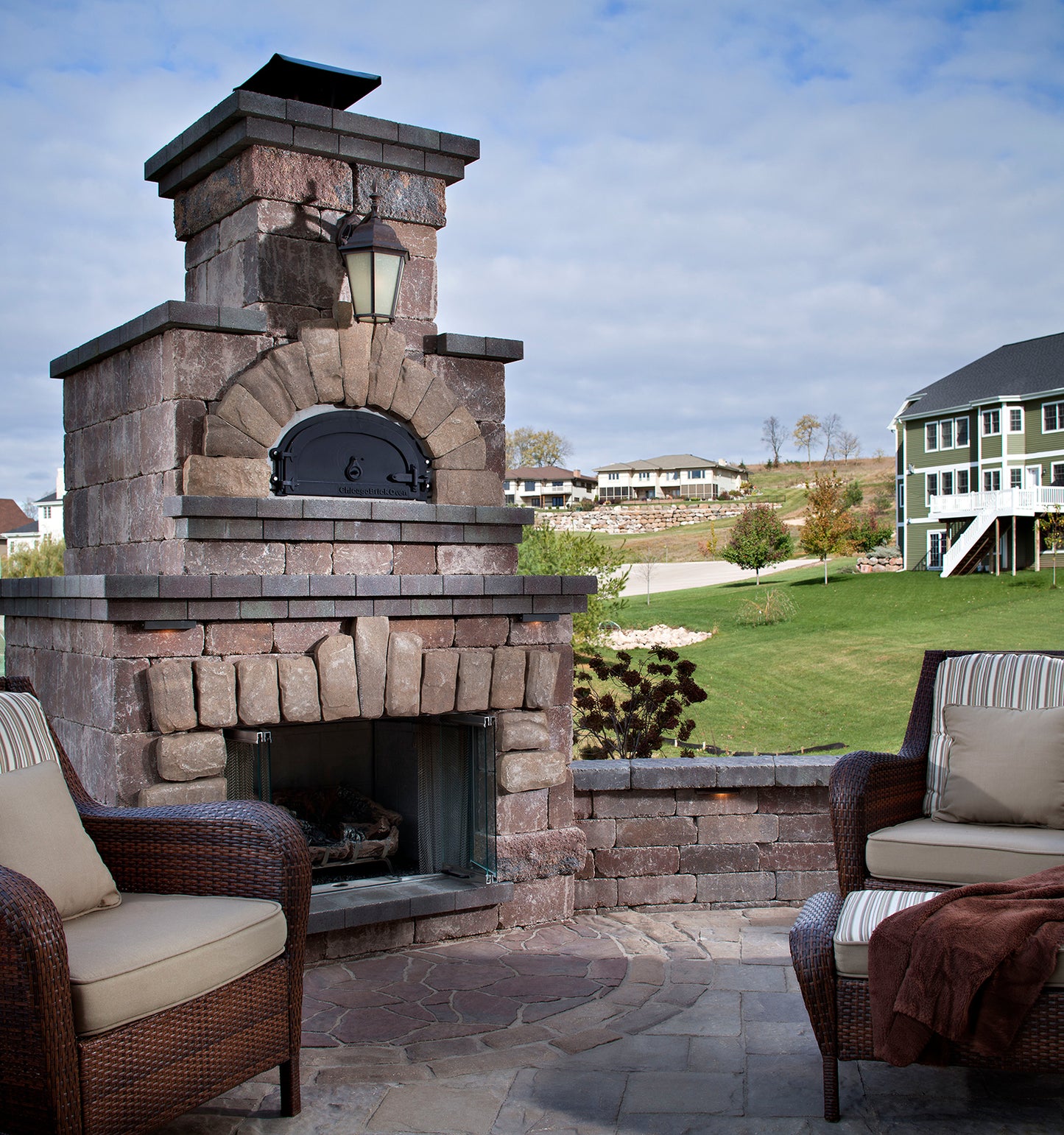 A patio scene features the CBO 750 DIY Kit by Chicago Brick Oven, a large stone outdoor fireplace that doubles as a high-performance wood-fired pizza oven with a 38" x 28" cooking surface. Two cushioned wicker chairs, one with a blanket draped over the arm, are arranged on the stone patio. In the background, a grassy yard and houses are visible under a partly cloudy sky.