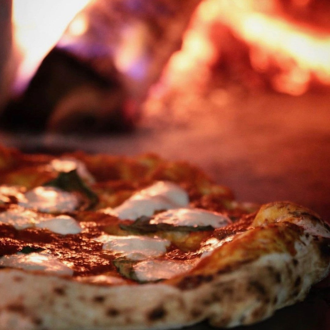 Close-up of a wood-fired pizza garnished with melted mozzarella cheese and tomato sauce. The pizza has a golden, slightly charred crust. In the background, there's an out-of-focus view of flames inside a Chicago Brick Oven CBO 750 Hybrid DIY Kit (Residential), made with refractory cement and offering both gas and wood fuel options, creating a warm ambiance.