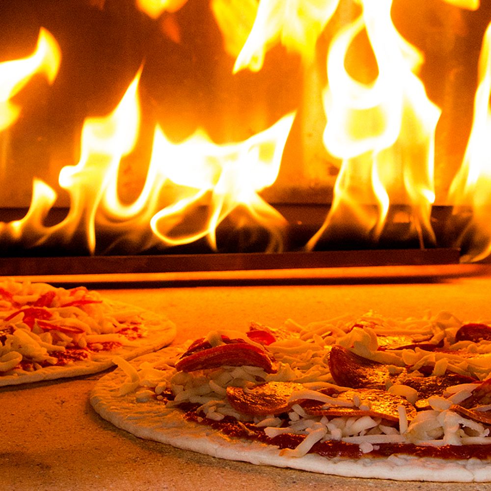 Two pizzas with toppings, including pepperoni and cheese, are baking in a Chicago Brick Oven CBO 750 Hybrid DIY Kit (Residential) | Dual-Fuel (Gas and Wood). The flames visible in the background of the wood-fired pizza oven provide an intense fire that casts a warm, orange glow, illuminating the pizzas and the refractory cement lining the oven’s interior.