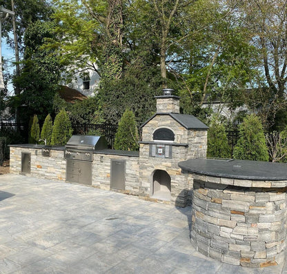 Outdoor kitchen with stone features, including the Chicago Brick Oven CBO 750 Hybrid DIY Kit (Residential) dual-fuel (gas and wood) pizza oven made of refractory cement, grill, and countertop. The kitchen is surrounded by lush greenery and trees, with a fence in the background. The area is paved with gray stone tiles.