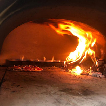 The CBO 750 Hybrid DIY Kit from Chicago Brick Oven, a dual-fuel outdoor oven that uses both gas and wood, is baking a freshly made pizza. Flames and glowing embers illuminate the pizza, highlighting the traditional cooking method. The pizza rests on a stone surface, encircled by refractory cement that lines the oven's curved interior for optimal heat distribution.