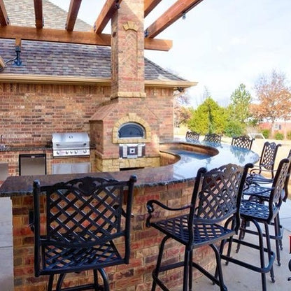 Outdoor kitchen with brick and stone construction featuring a Chicago Brick Oven CBO 750 Hybrid DIY Kit (Residential), which uses both gas and wood for firing, along with a grill and bar seating area. Four ornate metal bar stools align along the curved countertop, and a pergola with wooden beams provides partial shade. Trees are visible in the background.