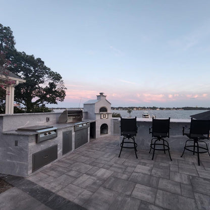 A spacious outdoor kitchen and dining area with a picturesque view overlooking a large body of water at sunset includes the Chicago Brick Oven CBO 750 Hybrid DIY Kit (Residential), which features a dual-fuel (gas and wood) built-in grill, wood-fired pizza oven with refractory cement. The setup also boasts ample countertop space, bar seating with four chairs, and views of trees and a distant shoreline.