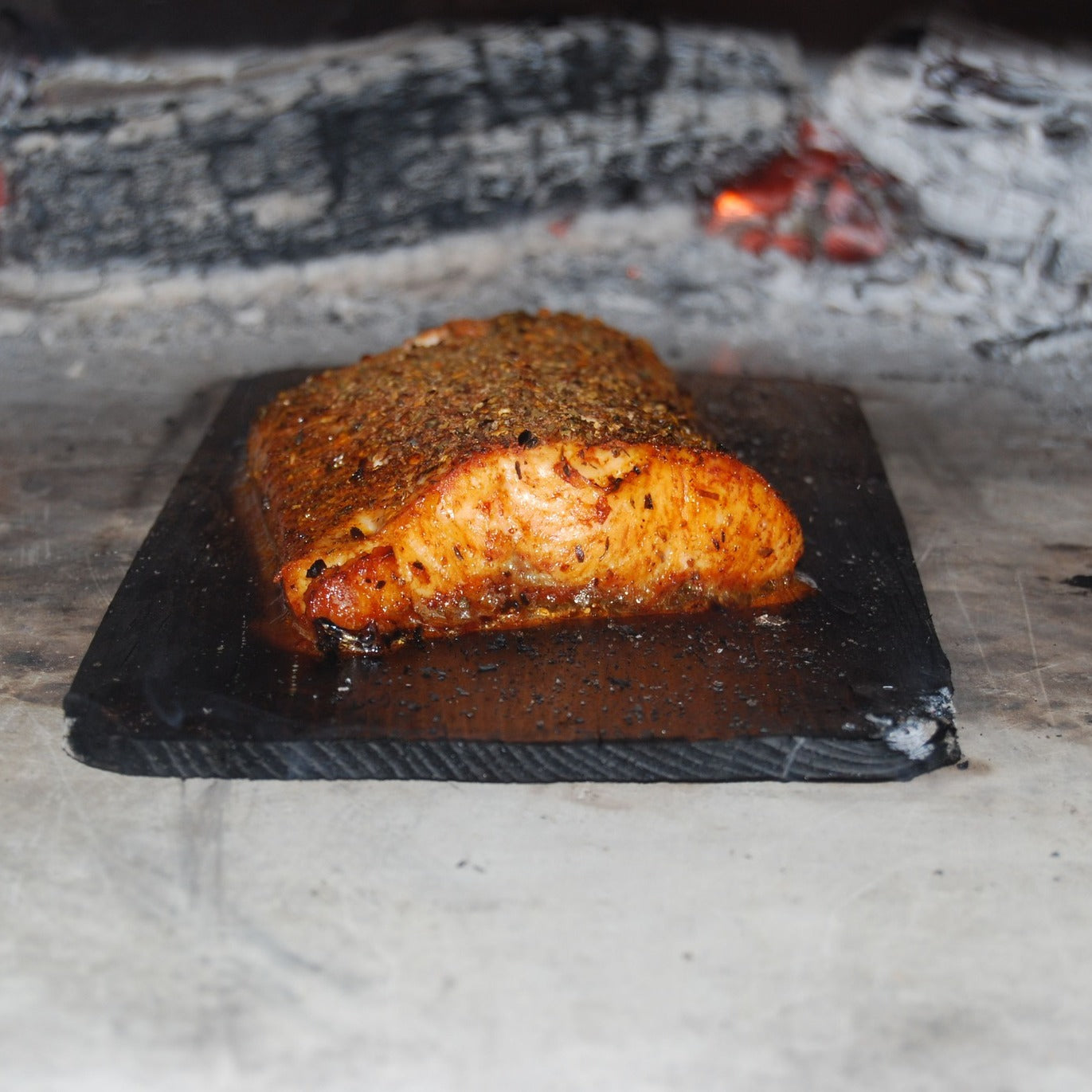 A seasoned salmon fillet is being cooked on a plank of wood, with a smoldering fire and charred logs from the Chicago Brick Oven CBO 750 Hybrid DIY Kit (Residential) | Dual-Fuel (Gas and Wood) in the background. The fish is golden brown, grilled to perfection with visible seasoning, and boasts a glistening surface.