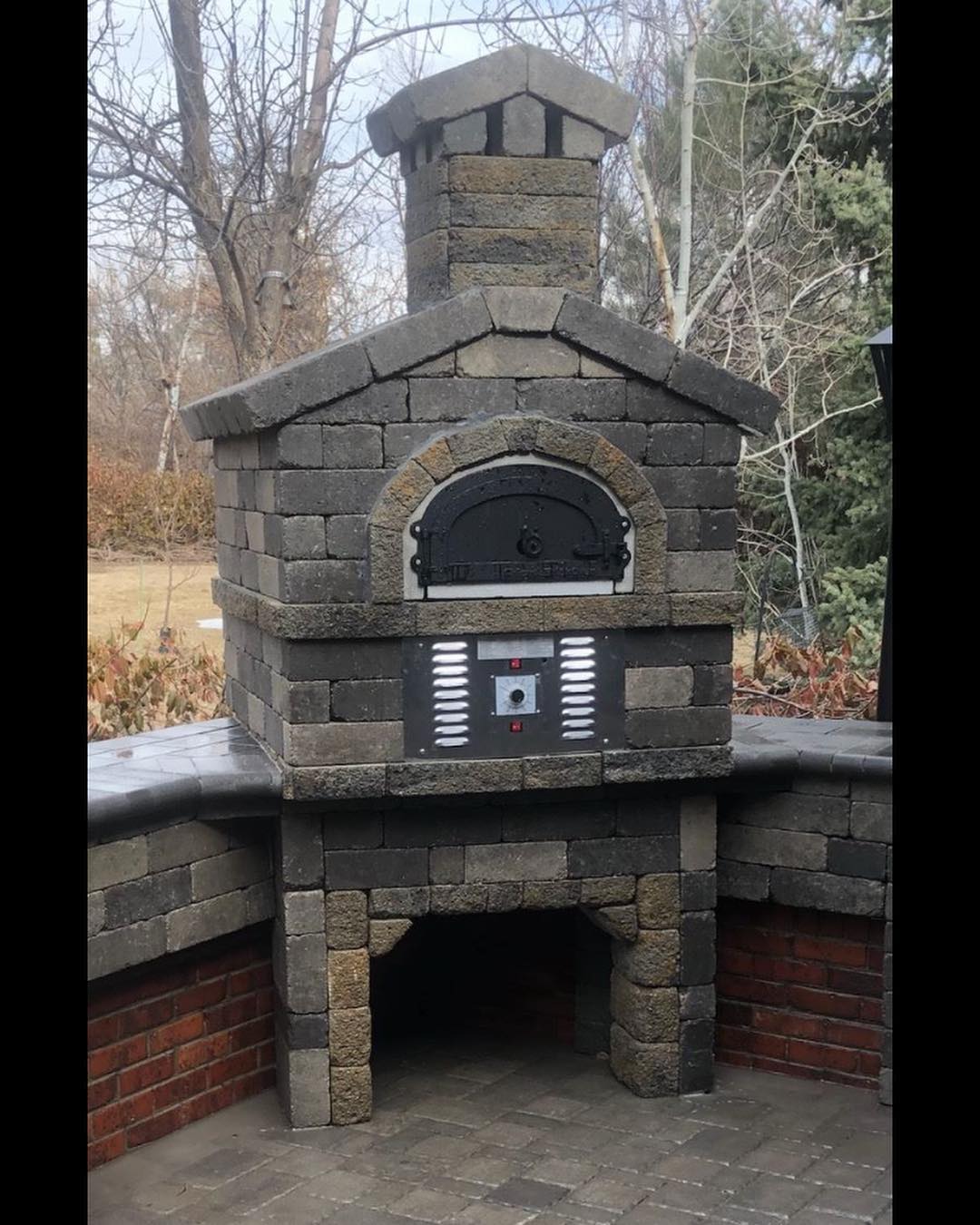 The CBO 750 Hybrid DIY Kit (Residential) by Chicago Brick Oven, a dual-fuel (gas and wood) outdoor pizza oven, stands on a stone platform. Built with refractory cement, it features a small chimney on top, a black metal door with a thermometer, and a lower storage area. The oven is surrounded by trees and bushes.