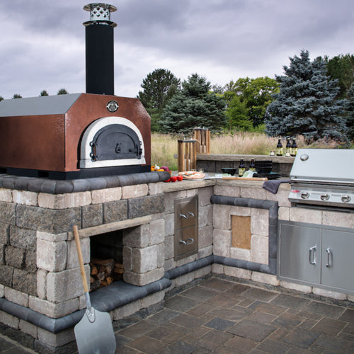 An outdoor kitchen featuring the Chicago Brick Oven's CBO 500 Countertop wood-fired pizza oven, with a chimney and a stainless steel grill. The stone countertops showcase various kitchen utensils, fresh vegetables, and a cutting board. A pizza peel leans against the commercial-grade refractory cement oven that boasts a 27" x 22" cooking surface, and the area is surrounded by trees.