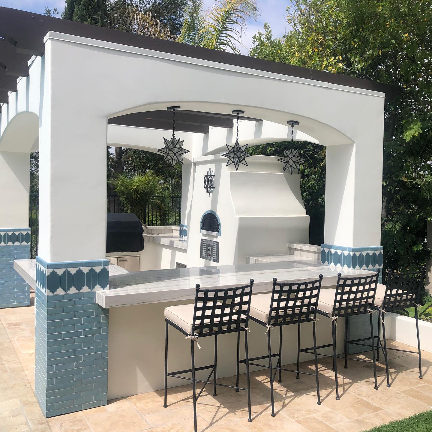 An outdoor kitchen showcases a white stucco structure with blue tile accents, featuring a built-in grill and the CBO 750 Hybrid DIY Kit (Residential) from Chicago Brick Oven, which offers dual-fuel options for both gas and wood. The setup includes three pendant lights, a high counter with three barstools, and is beautifully surrounded by greenery.