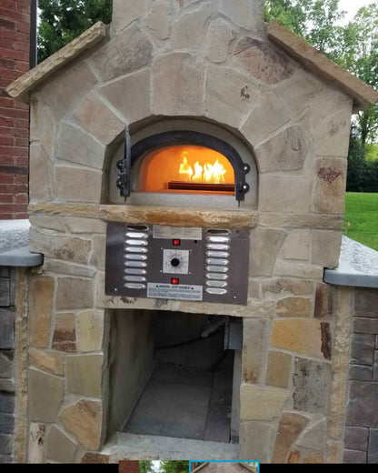 The CBO 750 Hybrid DIY Kit (Residential) by Chicago Brick Oven, a dual-fuel oven for both gas and wood, is crafted with refractory cement and features a metal control panel beneath the door along with a storage area underneath. Surrounded by brick and stone, it brings rustic charm to your outdoor space, complemented by lush greenery in the background.