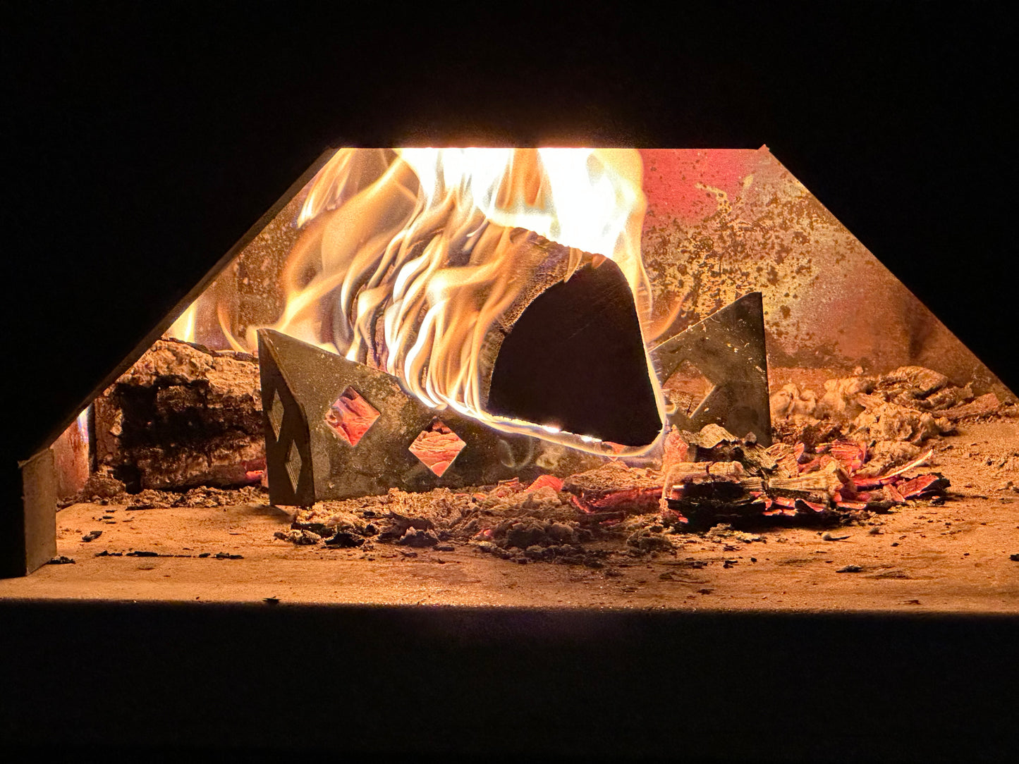 A close-up view of a burning log inside a Chicago Brick Oven. Resting on a premium Chicago Brick Oven Steel Log Holder, the log is positioned on a metal grate, with bright flames and glowing embers visible. The scene is warm and rustic, capturing the essence of a cozy, traditional fire.