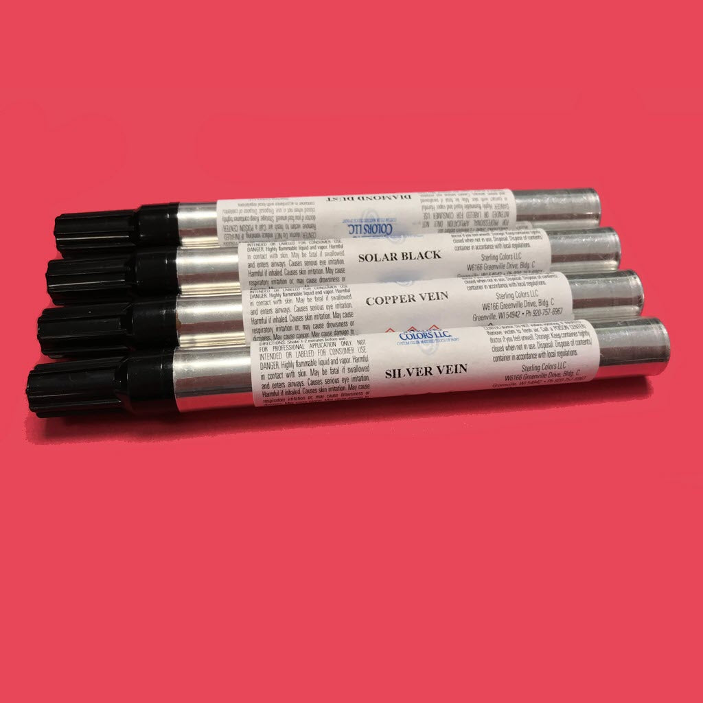 Four metallic paint markers with black caps are arranged in a fan-like pattern on a red background. Ideal for touch-up paint tasks, each marker from the Chicago Brick Oven Touch-Up Paint Kits is labeled with different colors: Solar Black, Copper Vein, and Silver Vein.