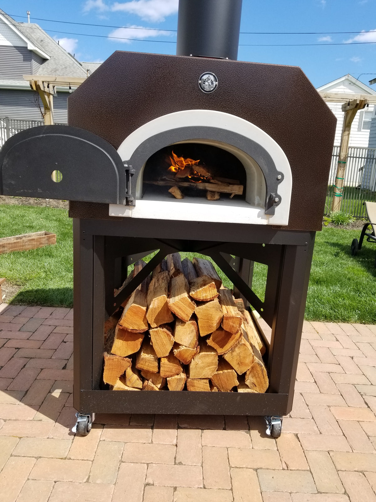 Copper Vein 750 Mobile oven with visible fire and firewood storage on a sunny brick patio
