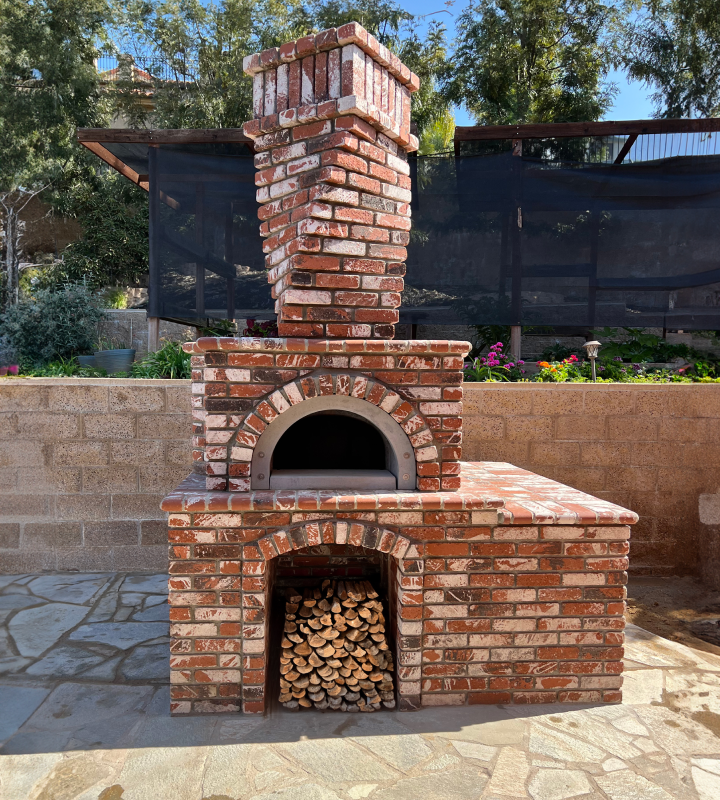 A brick outdoor pizza oven stands on a paved surface with a pile of firewood neatly stacked underneath. The oven features a tall chimney and is set against a background of greenery and a stone wall.