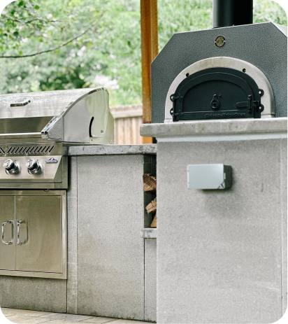 A modern outdoor kitchen featuring a stainless steel grill on the left and the Chicago Brick Oven CBO 500 Countertop wood-fired pizza oven with a 27" x 22" cooking surface crafted from commercial-grade refractory cement on the right. The light-colored stone countertop adds an elegant touch, while a small pile of firewood is neatly stacked underneath the pizza oven. Trees and a wooden fence provide a picturesque backdrop.