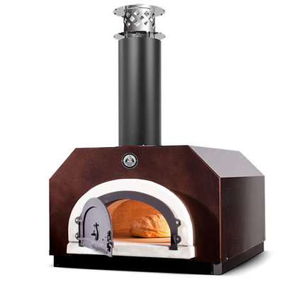 The CBO 500 Countertop Wood Fired Pizza Oven by Chicago Brick Oven features a compact, metal design with a visible chimney stack and is adorned in a striking dark brown finish. The outdoor wood-burning oven's front door stands open, revealing a loaf of bread baking inside, illuminated by the warm, golden glow from the fire within its 27" x 22" cooking surface.